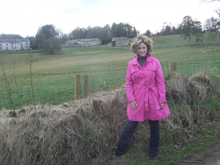 woman standing in a pink coat next to a hedge in rural ireland with gray buildings in background