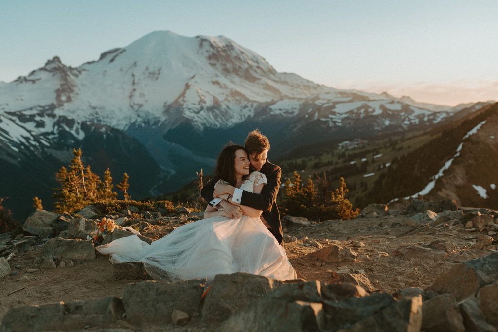Man in black suit, woman in white wedding gown sitting on the ground with snowcapped mountain in background.