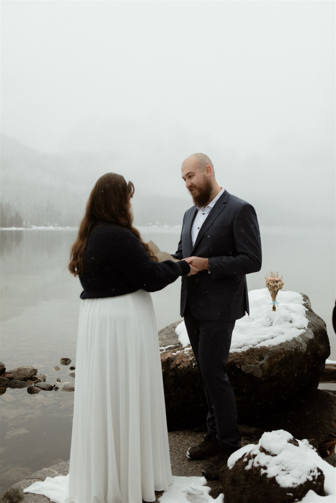 couple holding hands, sharing vows on lake shore in snow.