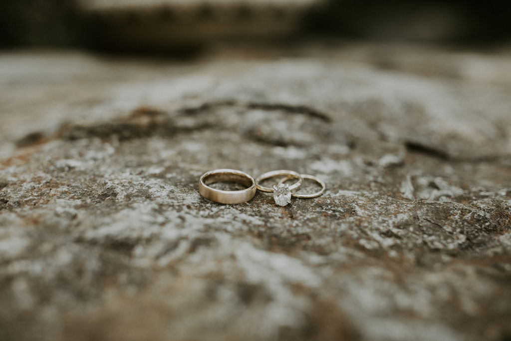 gold wedding bands and diamond solitaire engagement ring sitting on large rock. Photo courtesy of Greenhouse Photo Co.